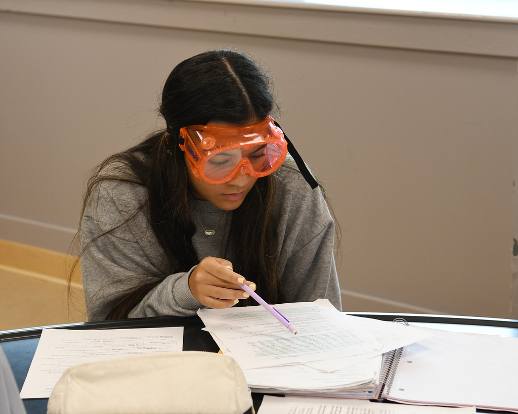 Person wearing orange safety goggles, seated at a table, studying a notebook and papers.
