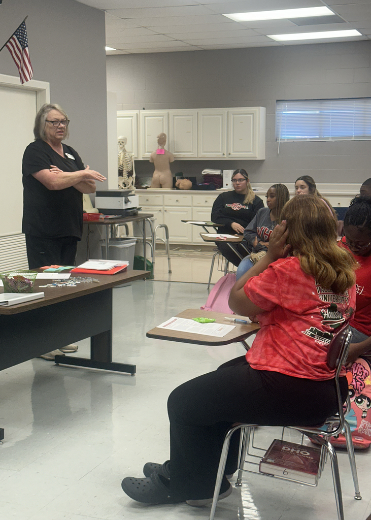 A woman, wearing glasses, teaches a seated class. The class has desks and a window. An American flag is visible.