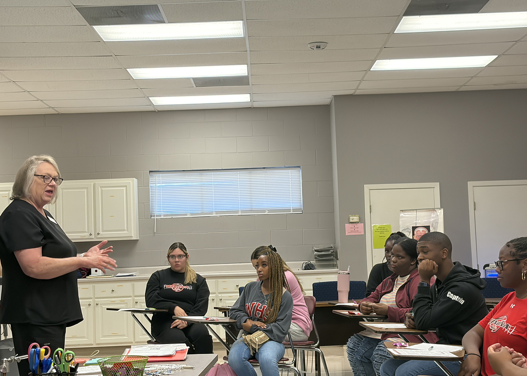 Instructor stands in front of students, speaking. Desks, chairs, and overhead lighting fill the room. A window provides natural light.