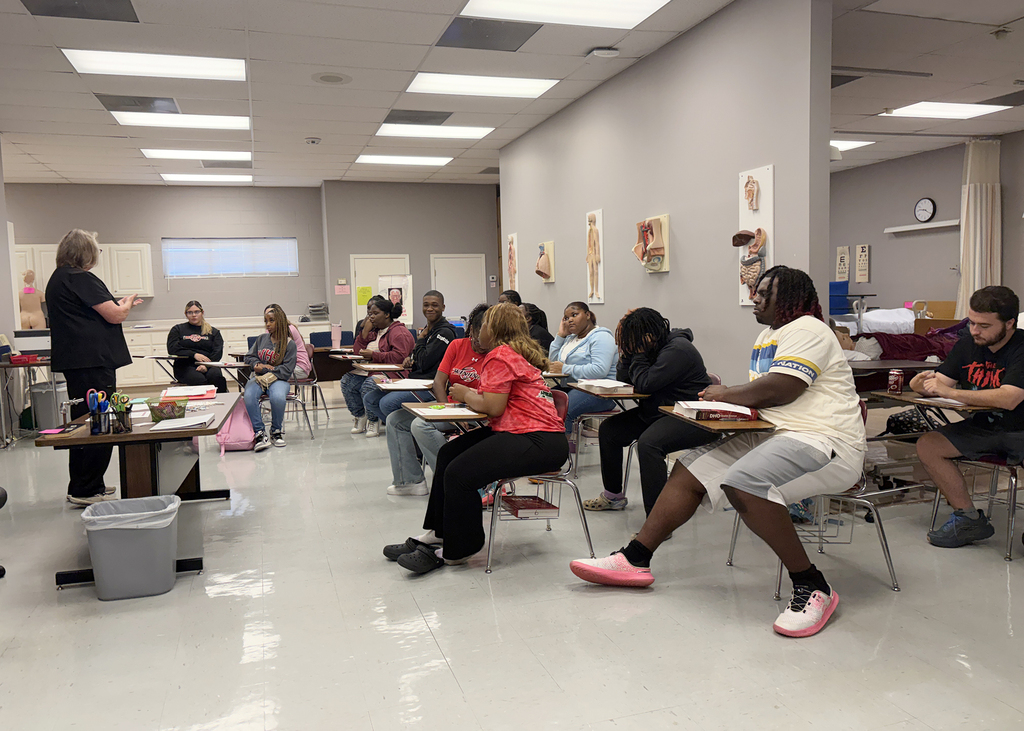 A classroom with white walls, ceiling lights, and a whiteboard. Students sit at desks, listening to a teacher.