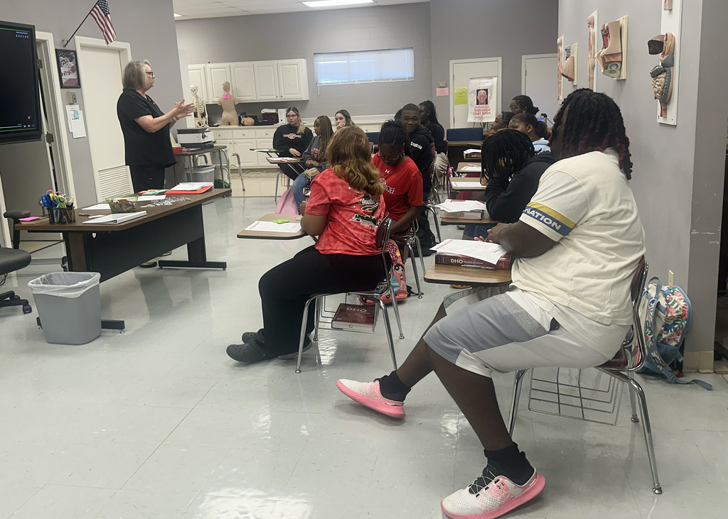 A classroom with students seated at desks, a teacher standing by a desk with a monitor, and a trash bin nearby.