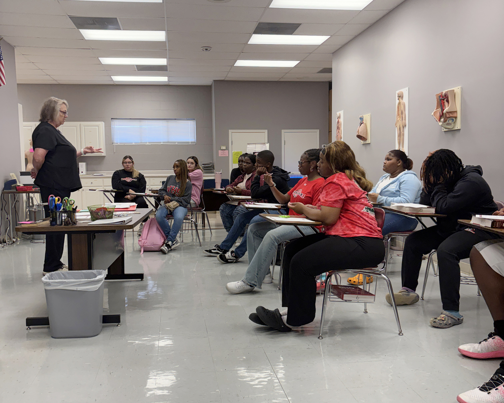 A classroom with gray walls has students sitting at desks. A woman stands at the front, and others are seated.