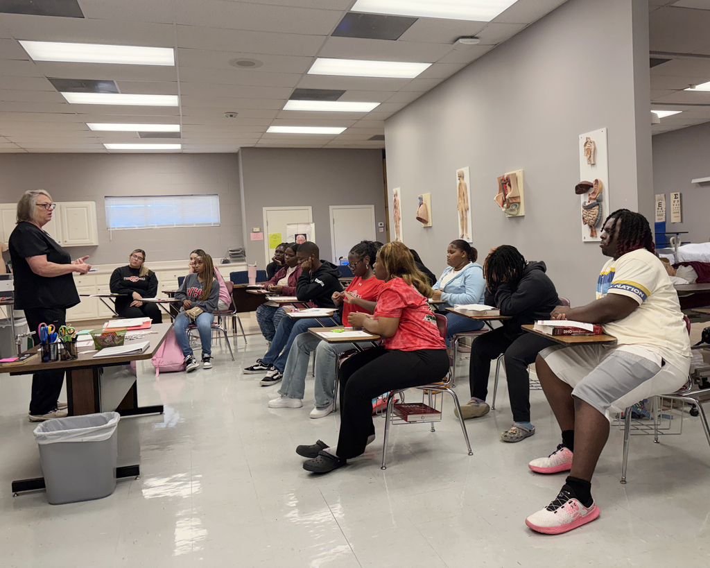 Classroom with an instructor and students seated at desks; walls with posters and gray flooring.