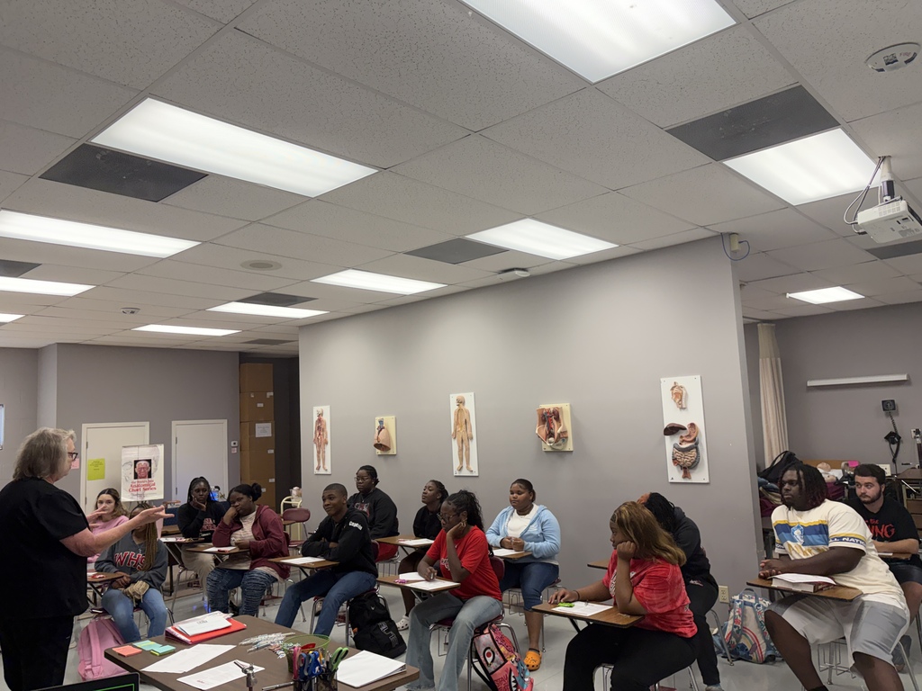 A classroom with a teacher in front, students seated in rows, tables, and posters on a wall.