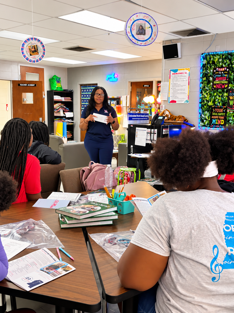 A classroom with a woman standing and speaking to students. Various books and papers are on the table.