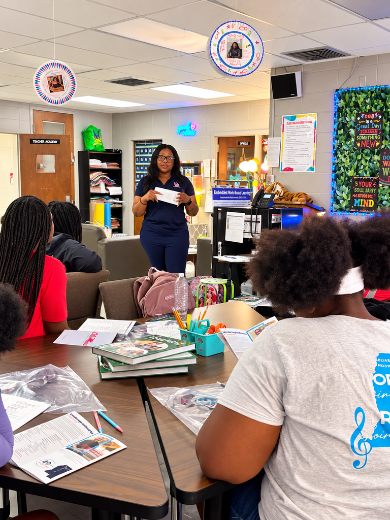 A teacher stands at a desk in a classroom, addressing students seated at tables. Classroom decorations adorn the walls.