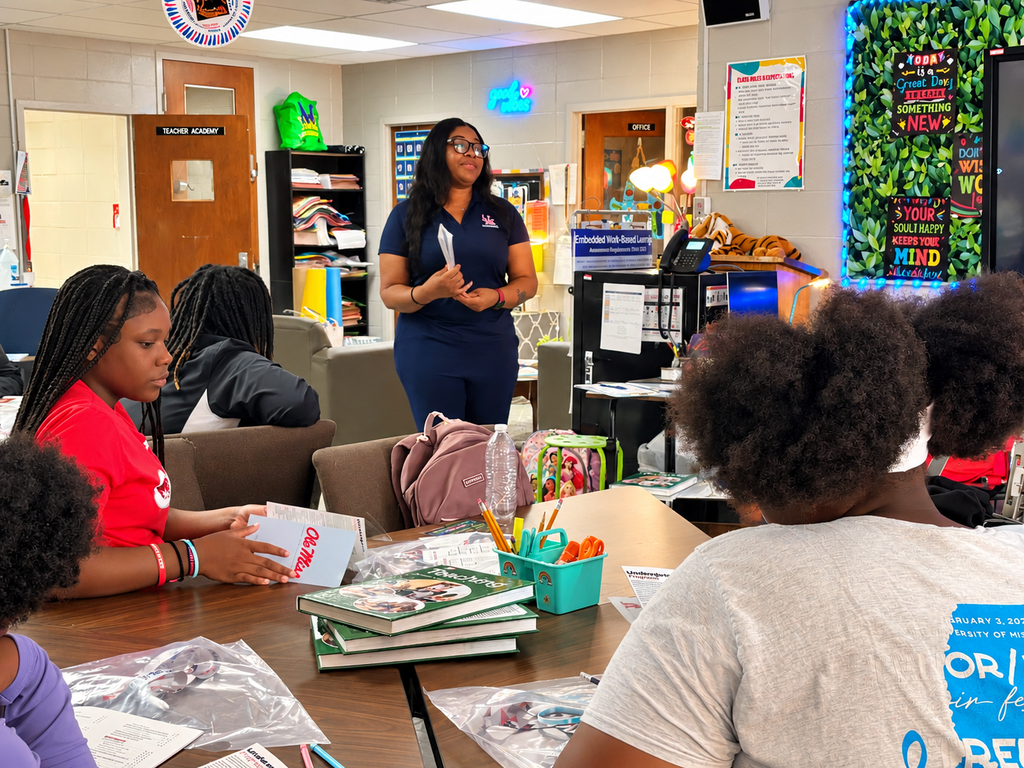 A woman stands before students in a classroom, leading a discussion. Students sit around a table with books and supplies.