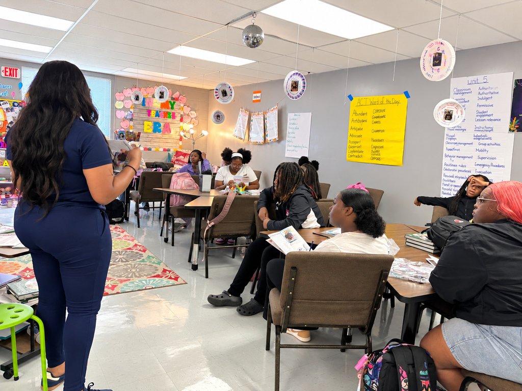 A woman stands in a classroom, addressing a group seated around tables. Papers and backpacks are on tables.