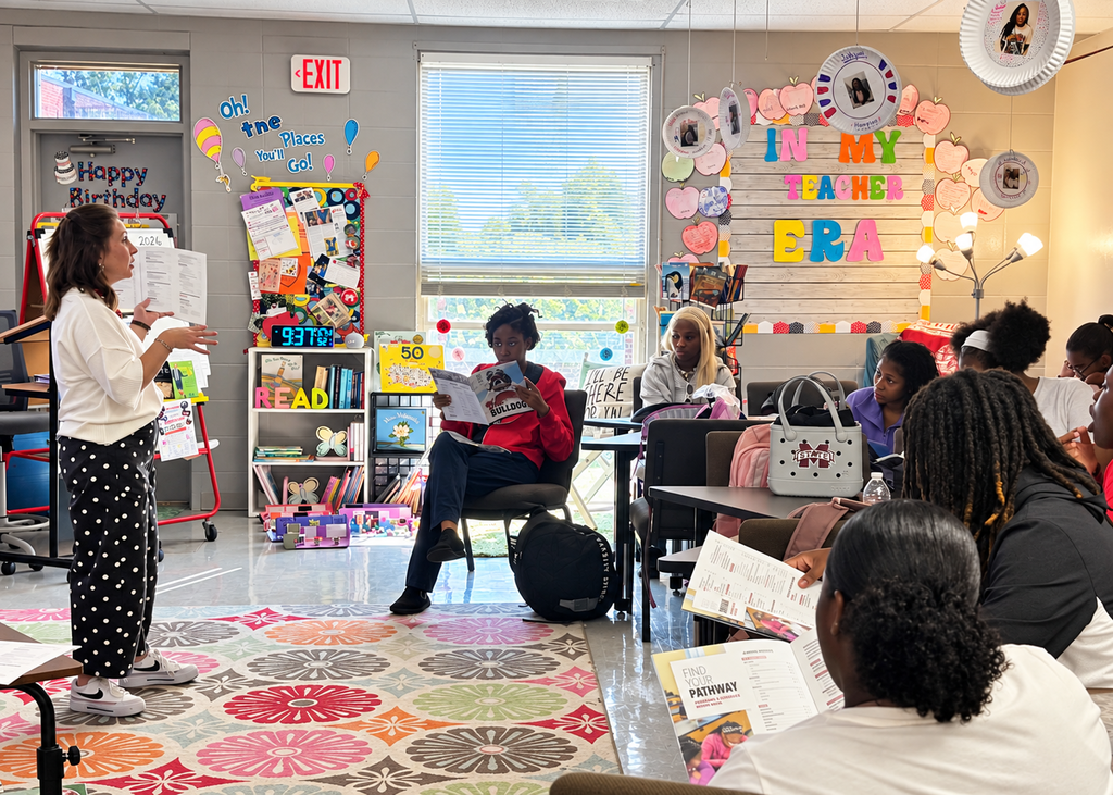 Teacher speaks to a group of students, one reading a book. Colorful decorations on walls and rug.