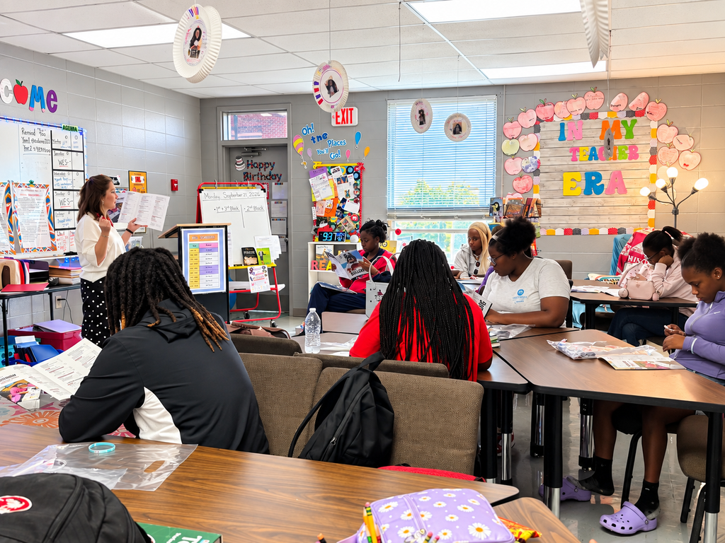 A classroom with desks and chairs. A woman stands in front of a whiteboard, speaking to students.