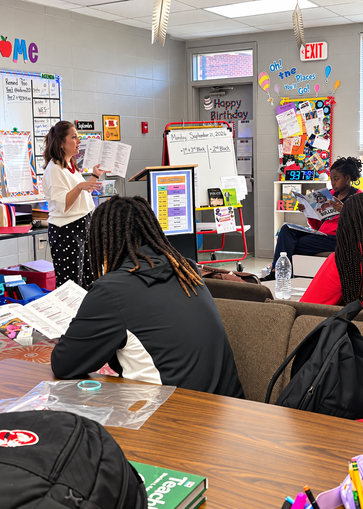 A teacher stands reading aloud in a classroom. Students are seated and listening. Brightly colored decorations adorn the walls.