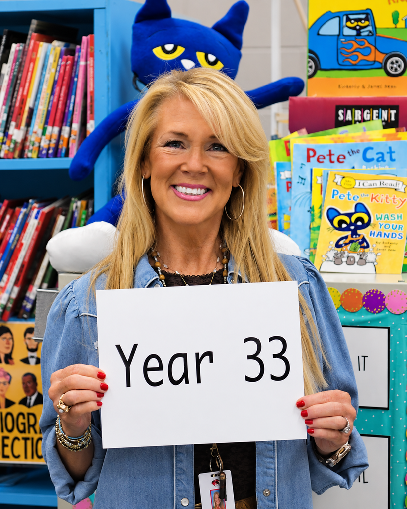 A woman holds a sign that reads "Year 33" in front of a colorful classroom with books and a large stuffed animal.