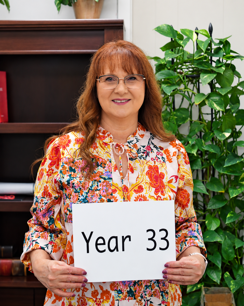 Woman holding a sign reading "Year 33" in front of a plant and a bookshelf.