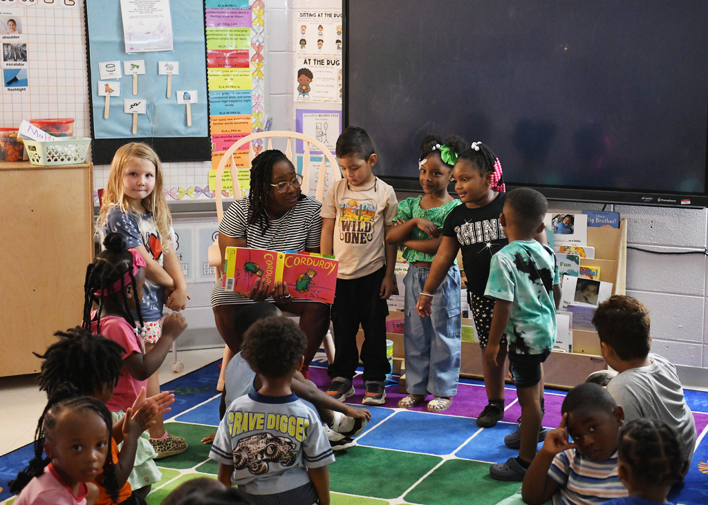 A classroom with a teacher reading a book to young children in front of a large screen.