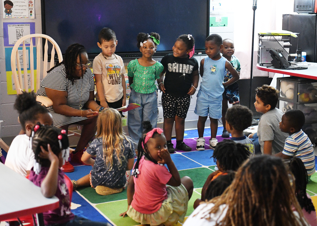 A classroom with a woman and young children sitting and standing on a colorful mat, facing a screen.