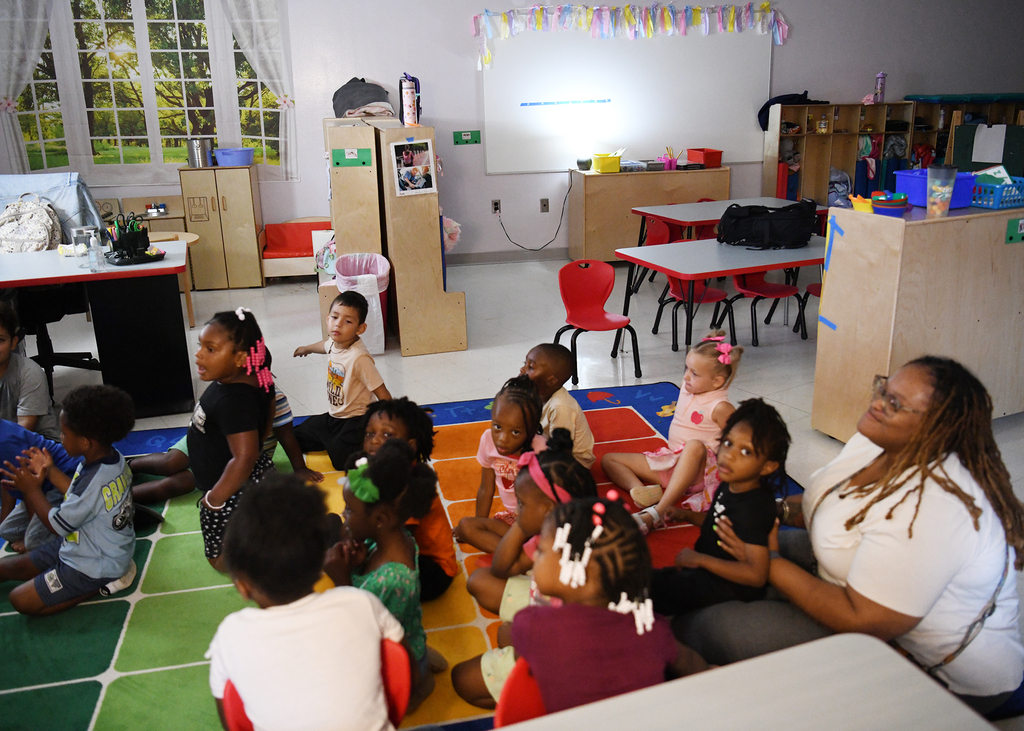 A classroom with children sitting in a circle, a woman with glasses sits cross-legged among them.