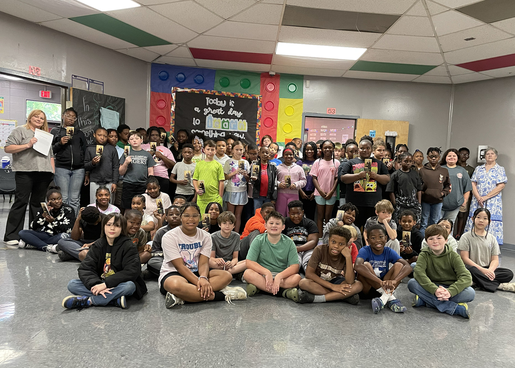 A large group of students seated and standing in a classroom. A teacher holds a book.