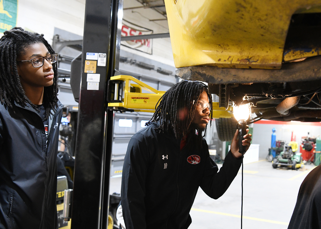 Two individuals in a garage; one examines a vehicle's underside with a flashlight while another stands nearby.