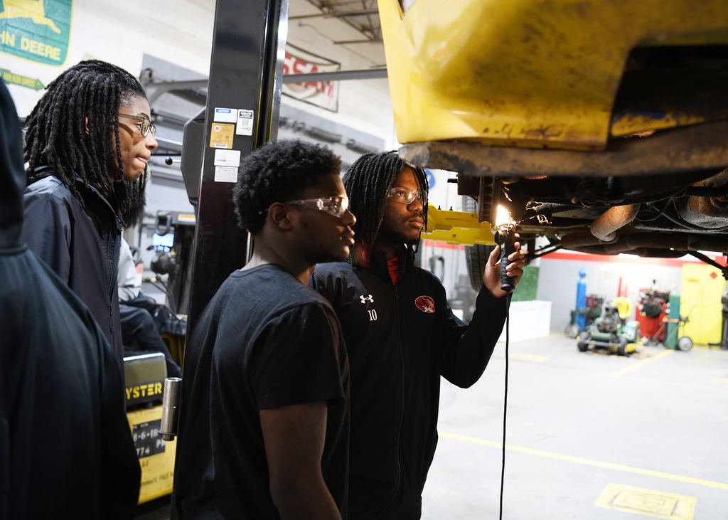 Four people in a garage, two are observing a vehicle on a lift with a yellow underside.