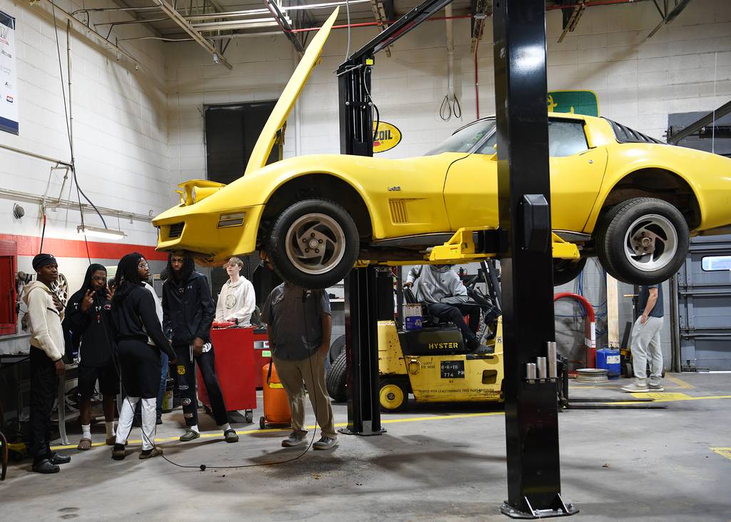 A yellow car is lifted by a jack in a shop. People stand around it. A forklift is parked nearby.