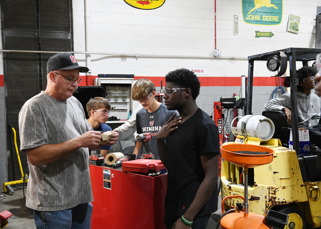 A man in a cap and eyeglasses speaks with a young man in a black shirt. Behind them, two other people stand. A forklift is to the right.