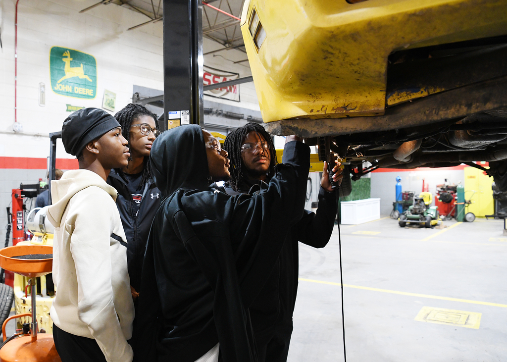 Four people in a garage, examining a yellow vehicle on a lift. The garage has tools and machines.