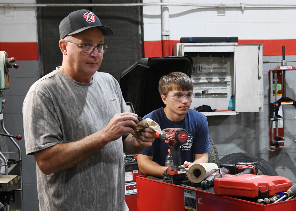 A man in a gray shirt and a cap stands in a garage while another man in a blue shirt works.