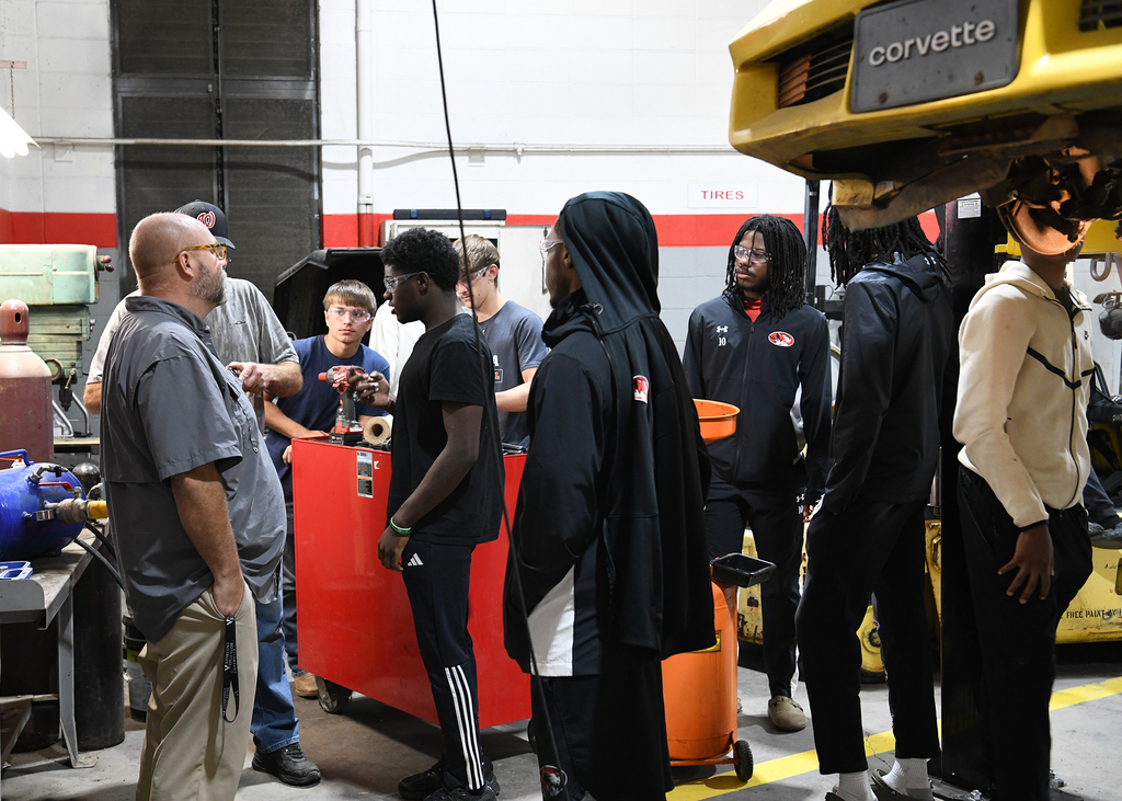 Group of people in a workshop with a yellow vehicle. Some are wearing jackets and sneakers.