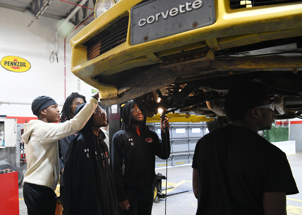 A group of people examine a yellow Corvette in a workshop. A man holds a flashlight.