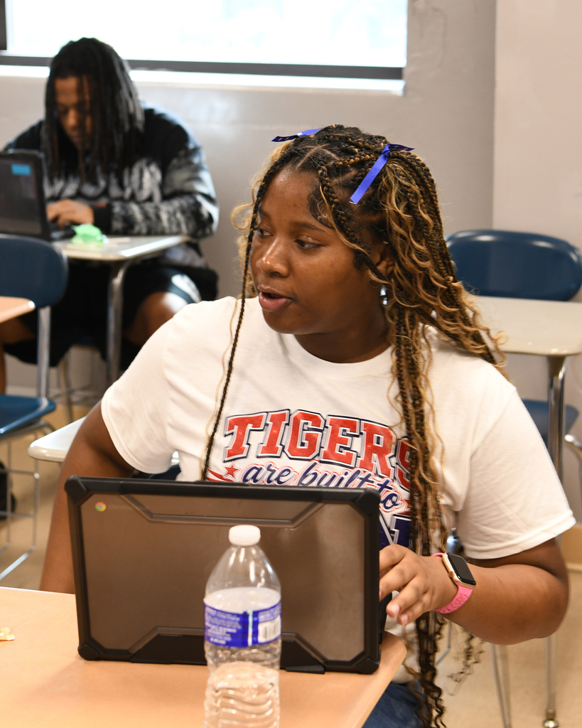 Person with long hair in a classroom sits at a table with a laptop and water bottle.