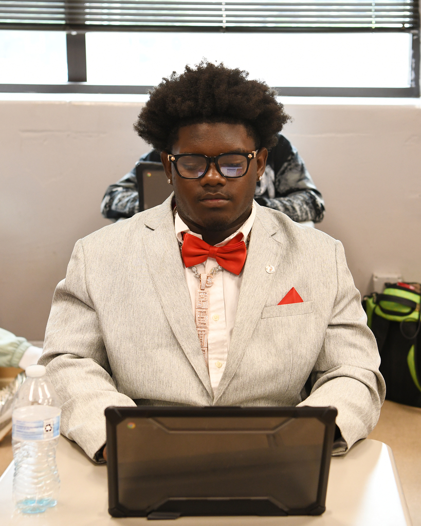 Person in gray suit with red bowtie seated at table using laptop. A water bottle and bag behind.