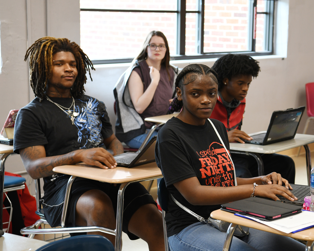 A classroom with several students seated at desks, engaged with laptops. One male and two females in front, others behind.