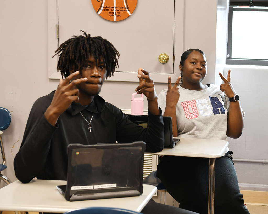 Two students sitting at desks, one gesturing peace sign, other using a laptop, basketball emblem on wall.