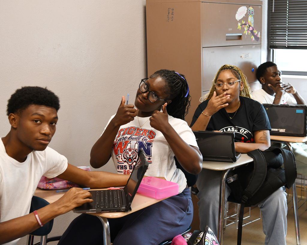 Four students in a classroom, seated at desks. One is using a laptop, and another holds a pen.
