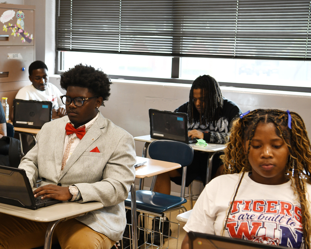 People using laptops in a classroom. One wears a bow tie, another has pigtails. Background includes blinds.