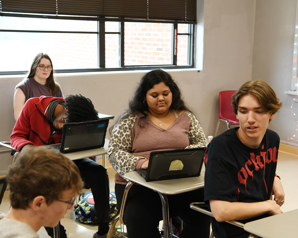 Students in a classroom with desks, laptops, and windows. Four individuals seated, each using a laptop.