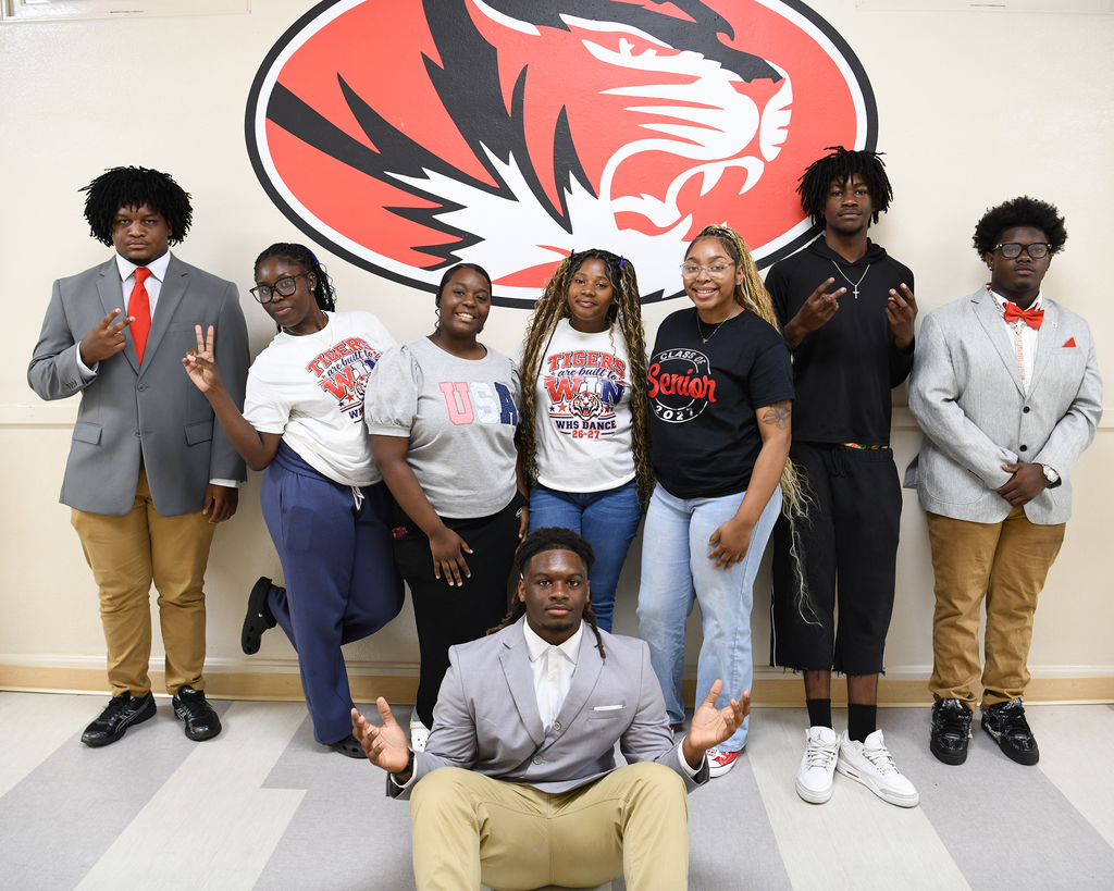 Eight people pose for a photo with a tiger emblem in the background. Some wear suits and sneakers.