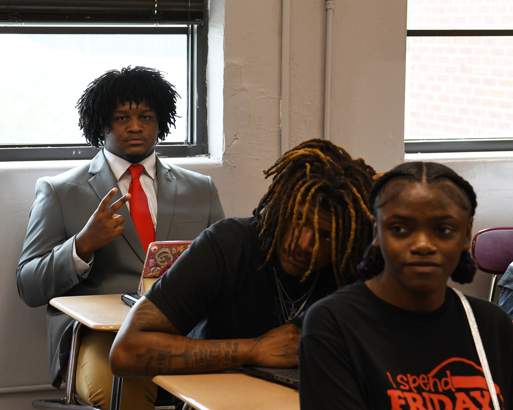 Three people in a classroom setting; one man with a red tie, another with dreadlocks, and a woman in a black shirt.
