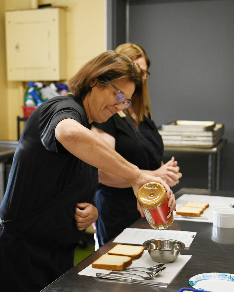 Two women in black shirts prepare food in a kitchen, one spreading peanut butter on bread.