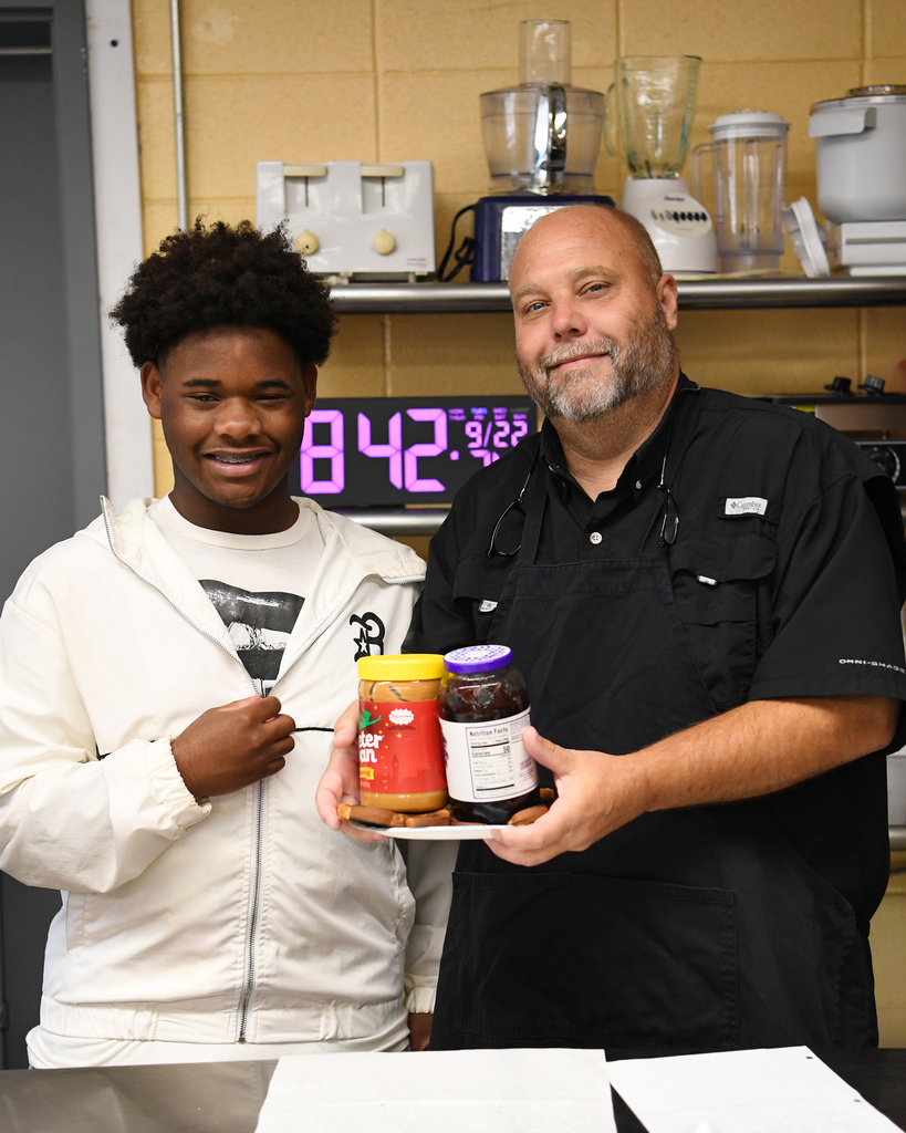 Two people in a kitchen; one holds a tray with three jars, the other holds a knife.