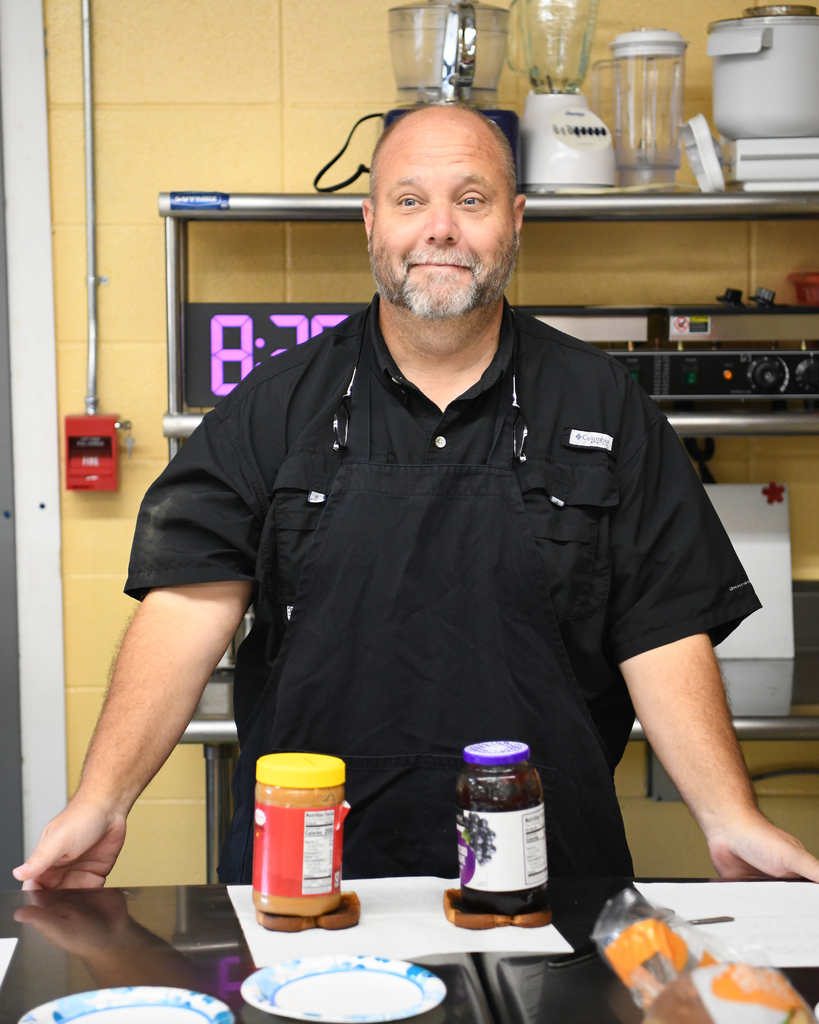 A man stands behind a counter in a kitchen. Two jars, one with peanut butter and another with jam, are on a table.