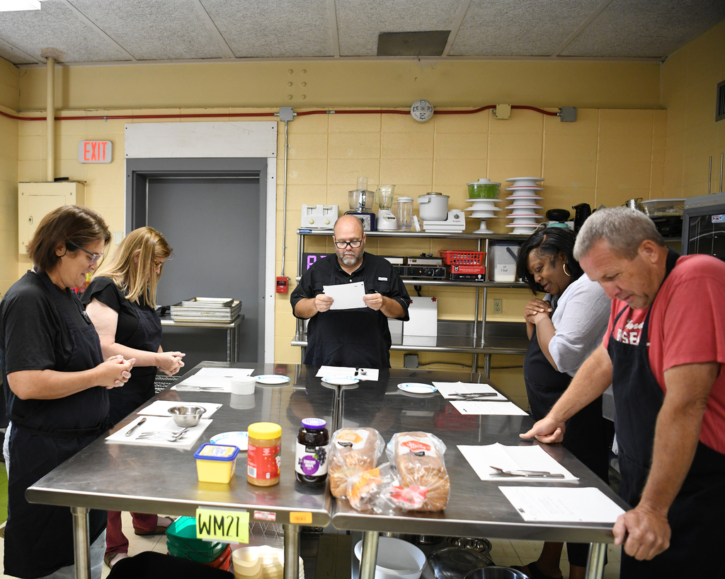 A group of people in aprons gather around a table with food items, led by a person with a paper.