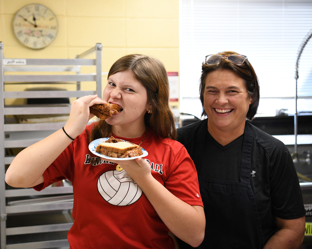 A woman eating a sandwich, another woman smiling beside her. A clock and oven in the background.
