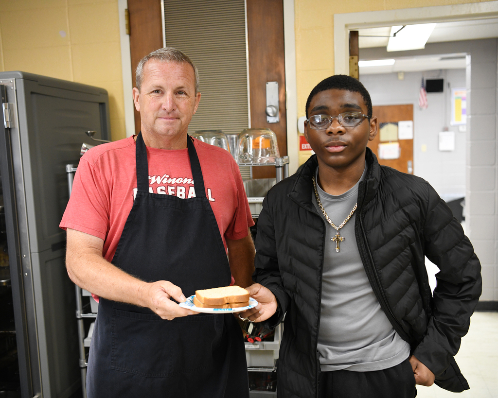 Two men in a kitchen. One holds a plate with a sandwich. Yellow walls, a metal cabinet, and a door.
