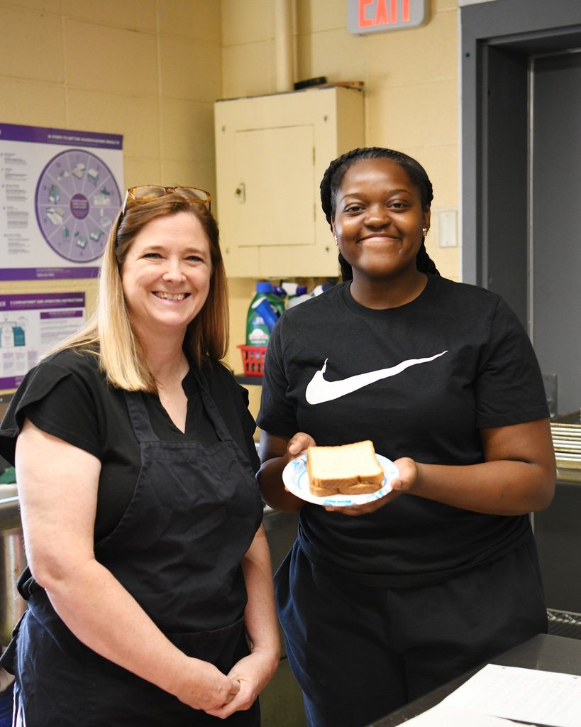 Two women, both in black uniforms, stand in a kitchen. One holds a sandwich. Behind them, a cabinet is on the wall.