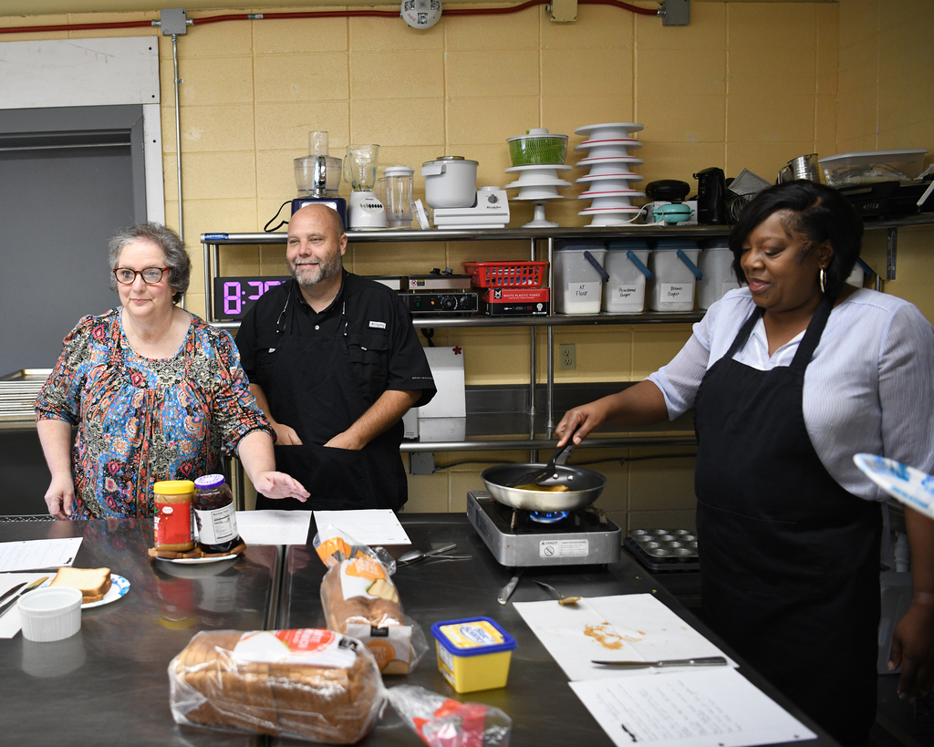 Three people in a kitchen. One holds a pan, another holds a knife. Bread, jars, and papers on table.
