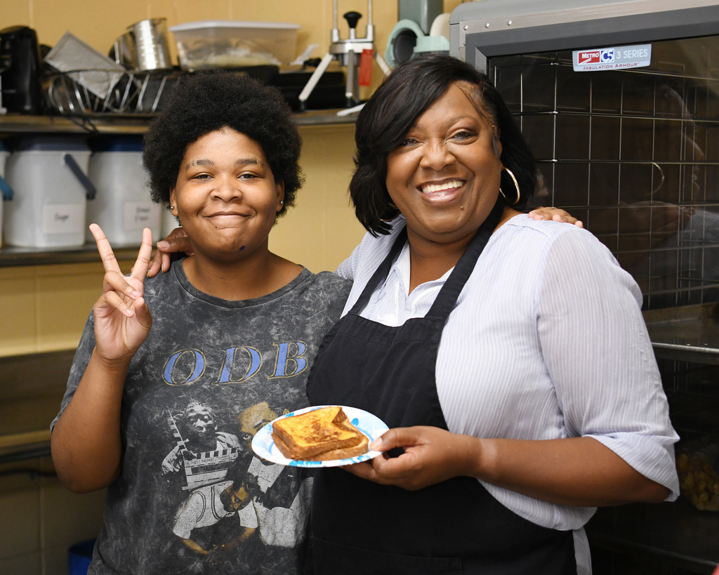 Two women in a kitchen, one holding a plate with food, smiling, and the other making a peace sign.