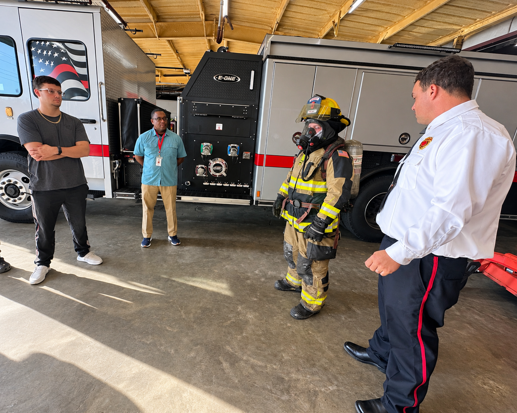 Four individuals, including a firefighter, in a fire station. One man speaks to the firefighter.