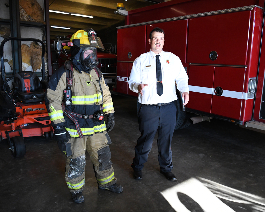A firefighter stands in front of a fire truck in a garage. The firefighter wears a mask.