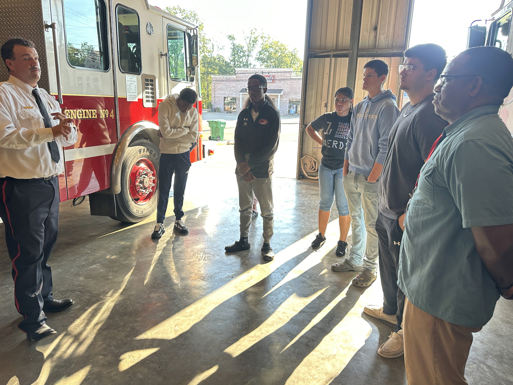 A group of people stands in a fire station with a fire truck. A man in white speaks to others.